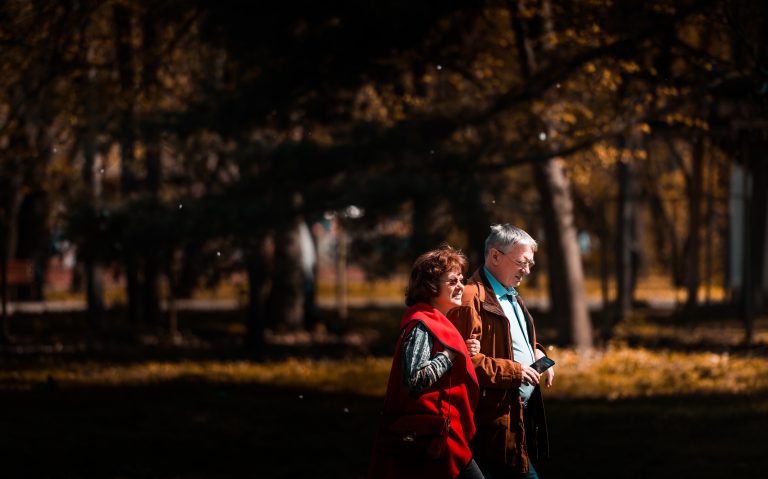 The image depicts an older couple having a walk in an autumn park. Both partners look energetic and full of life.