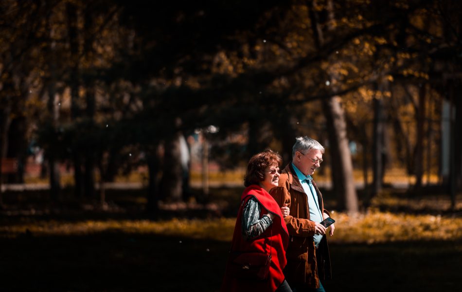 The image depicts an older couple having a walk in an autumn park. Both partners look energetic and full of life.