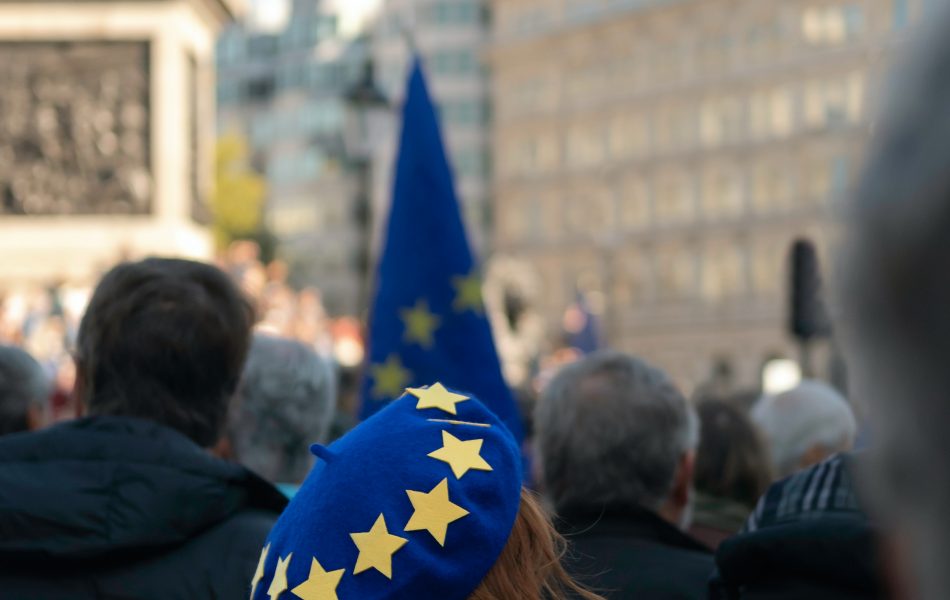 The image depicts a woman wearing a blue beret with yellow stars. The woman is a part of a street protest in some European city.